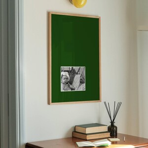 May include: A rectangular wall-mounted frame with a dark green mount featuring a small black and white photograph of a person in the centre. The frame is made of light-coloured wood and is positioned on a white wall below a yellow circular light.