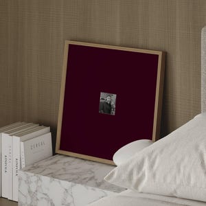 May include: A square picture frame with a light wood border and a deep burgundy mount. Centered within the mount is a small, rectangular black and white photograph depicting two individuals standing side by side in a portrait orientation.