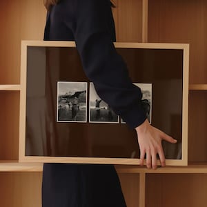 May include: A framed artwork with three black and white photographs. The frame is made of light-coloured wood. The photographs show scenes of people and horses, possibly from a Western or rural setting. The artwork is held up against a wooden shelf.