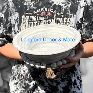 May include: A decorative, light gray bowl with a textured rim and a base adorned with wooden beads. The bowl has the text "Langford Decor & More" on the side. A natural fiber tassel hangs from the base. The bowl is held by a person.