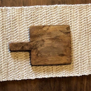 May include: A rustic wooden cutting board with a handle. The board is brown and has a natural wood grain. It is sitting on a white and brown woven rug.