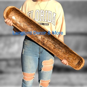 May include: A long, hand-carved wooden dough bowl with a natural, rustic finish. The bowl is held by a person wearing a yellow t-shirt and ripped blue jeans. The bowl's dimensions are approximately 30 inches long.