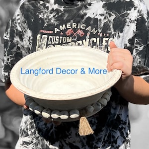 Puede incluir: Un cuenco decorativo grande, blanquecino, con una base de cuentas y una borla de fibra natural. El cuenco lo sostiene una persona que lleva una camisa teñida con tinte anudado en blanco y negro con el texto "AMERICAN CUSTOM CYCLES". El texto "Langford Decor & More" está impreso en el cuenco.