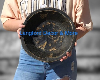 May include: A large, round wooden bowl with a dark brown stain and a distressed, aged look. The bowl is held by a person wearing blue jeans.
