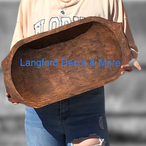 May include: A large, rustic wooden dough bowl with a natural brown finish. The bowl is oval-shaped and has a slightly raised rim. The bowl is being held by a person, but the person's hands are not visible in the image. The text "Langford Decor & More" is visible in the image.