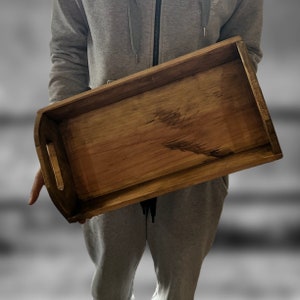 Może przedstawiać: Drewniana taca do serwowania z naturalnym wykończeniem drewna. Taca ma kształt prostokąta ze zaokrąglonymi krawędziami i podniesionym rantem wokół obwodu. Taca ma uchwyt po każdej stronie.