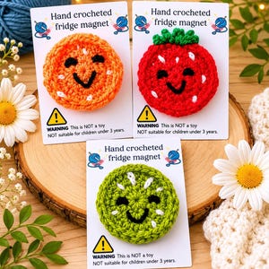 May include: Three hand-crocheted fridge magnets shaped like fruit slices: an orange, a strawberry, and a kiwi. Each features a smiling face, white embroidered seeds, and is attached to a white card labelled Hand crocheted fridge magnet.