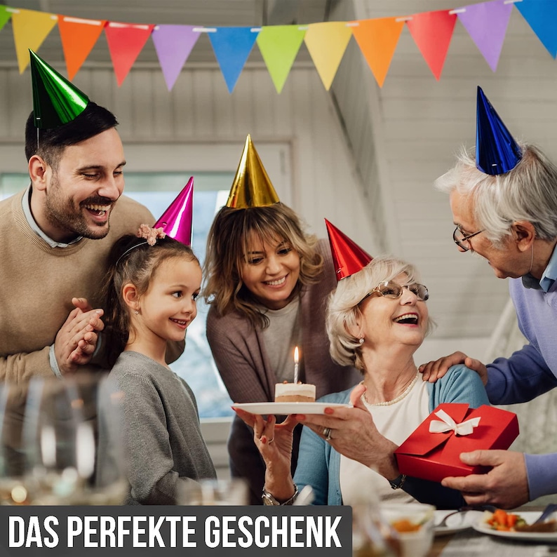 May include: A family celebrates a birthday with a small cake and a gift. The image shows a multi-generational group wearing party hats and smiling. A banner of colorful flags hangs in the background. The text at the bottom reads "DAS PERFEKTE GESCHENK".