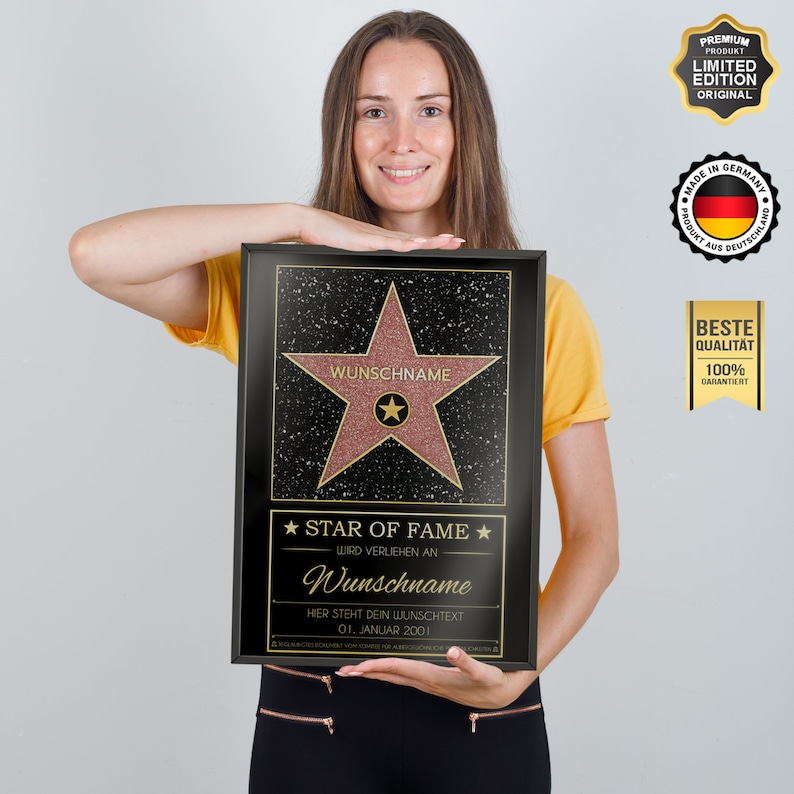 May include: A black plaque with a gold star and the text "STAR OF FAME" and "WIRD VERLIEHN AN" in gold. The star is pink and has the text "WUNSCHNAME" in gold. The plaque also has the text "HIER STEHT DEIN WUNSCHTEXT" and "01. JANUAR 2001" in gold.