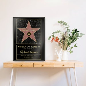 May include: A black framed poster with a pink star on a black background. The star has gold lettering that says "WUNSCHNAME" and "STAR OF FAME". The text below the star says "WIRD VERLIEHEN AN" and "Wunschname". The text below that says "HIER STEHT DEIN WUNSCHTEXT" and "01. JANUAR 2001".