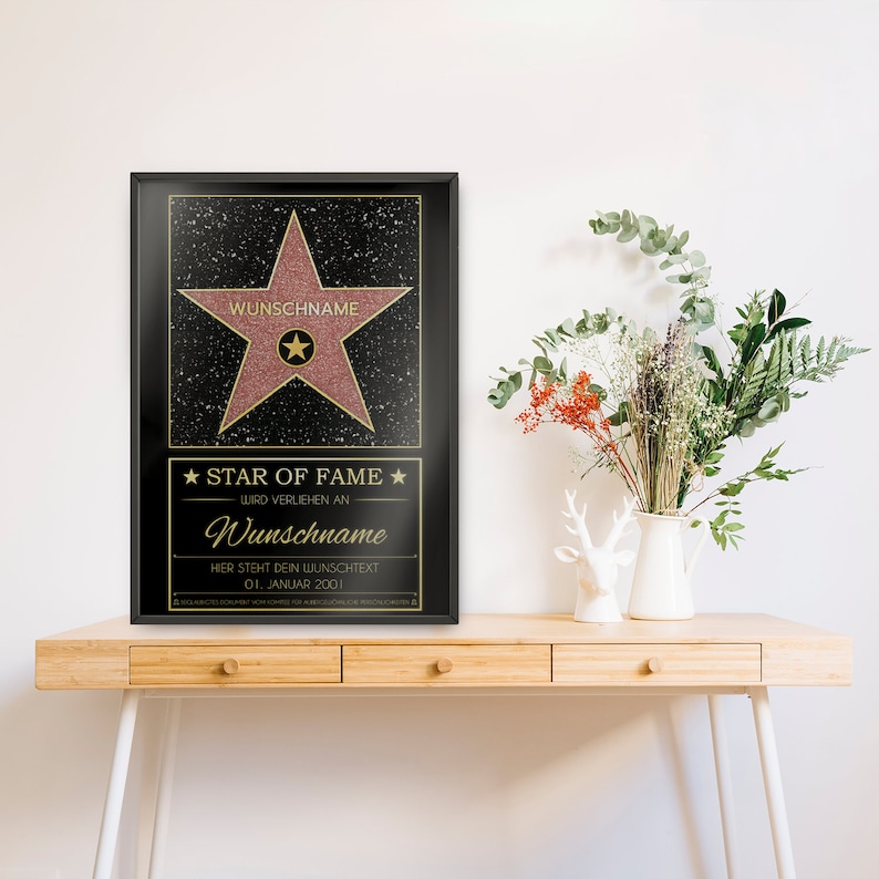May include: A black framed poster with a pink star on a black background. The star has gold lettering that says "WUNSCHNAME" and "STAR OF FAME". The text below the star says "WIRD VERLIEHEN AN" and "Wunschname". The text below that says "HIER STEHT DEIN WUNSCHTEXT" and "01. JANUAR 2001".