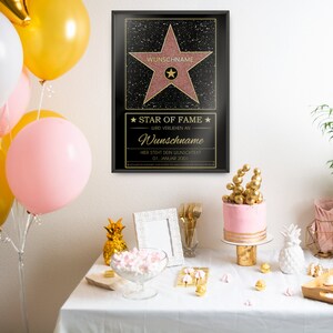 May include: A black framed poster with a gold star on a pink background. The poster says "Star of Fame" and "Wunschname" in gold lettering. The poster is on a table with a pink cake, gold pineapple decorations, and a white picture frame.