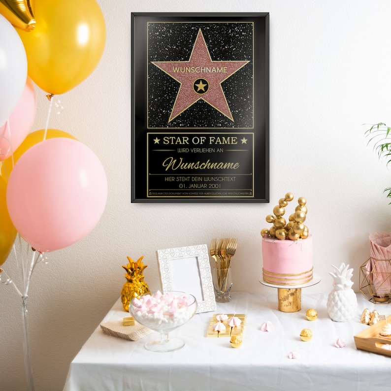 May include: A black framed poster with a gold star on a pink background. The poster says "Star of Fame" and "Wunschname" in gold lettering. The poster is on a table with a pink cake, gold pineapple decorations, and a white picture frame.