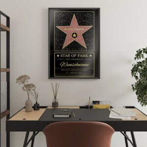 May include: A black framed poster with a gold star on a black background. The star has a pink center and the text "WUNSCHNAME" is written in gold at the top of the star. The text "STAR OF FAME" is written in gold at the bottom of the poster. The text "WIRD VERLIEHEN AN" is written in gold below the star. The text "Wunschname" is written in gold below the text "WIRD VERLIEHEN AN". The text "HIER STEHT DEIN WUNSCHTEXT" is written in gold below the text "Wunschname". The text "01. JANUAR 2001" is written in gold below the text "HIER STEHT DEIN WUNSCHTEXT".