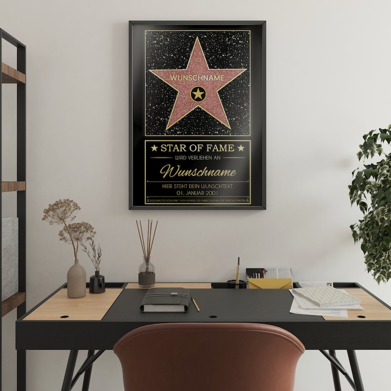 May include: A black framed poster with a gold star on a black background. The star has a pink center and the text "WUNSCHNAME" is written in gold at the top of the star. The text "STAR OF FAME" is written in gold at the bottom of the poster. The text "WIRD VERLIEHEN AN" is written in gold below the star. The text "Wunschname" is written in gold below the text "WIRD VERLIEHEN AN". The text "HIER STEHT DEIN WUNSCHTEXT" is written in gold below the text "Wunschname". The text "01. JANUAR 2001" is written in gold below the text "HIER STEHT DEIN WUNSCHTEXT".