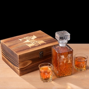 May include: A wooden box, a glass decanter, and two shot glasses are displayed on a wooden surface. The decanter and shot glasses are filled with a brown liquid. The box and decanter are engraved with the letter "M" and the name "Michael".