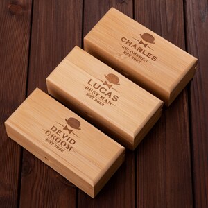 Puede incluir: Tres cajas de madera con texto grabado y un peque&ntilde;o dise&ntilde;o de corbata de lazo. Las cajas est&aacute;n etiquetadas "Charles - Groomsmen - Est 2022", "Lucas - Best Man - Est 2022" y "Devid - Groom - Est 2022".