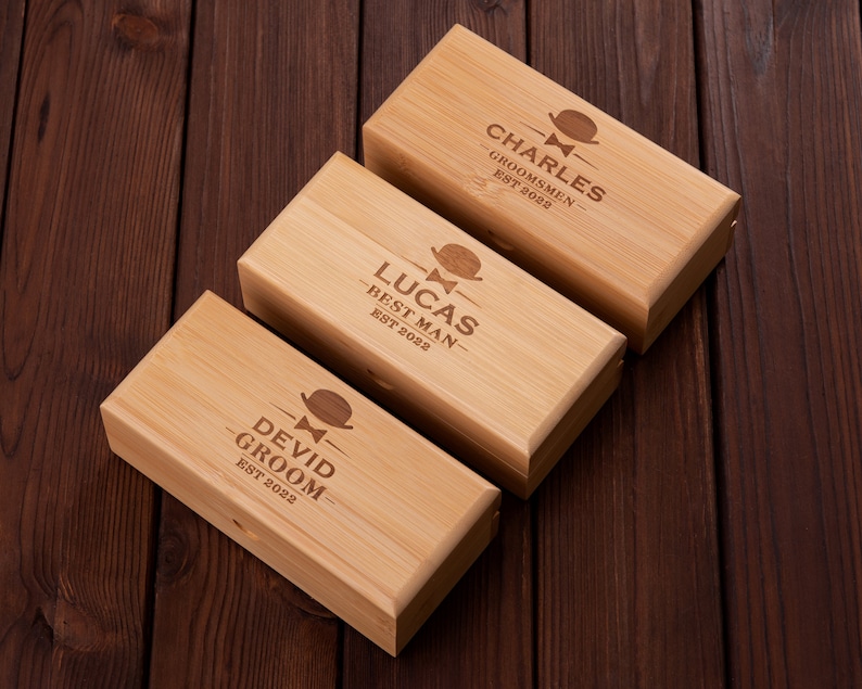 Puede incluir: Tres cajas de madera con texto grabado y un peque&ntilde;o dise&ntilde;o de corbata de lazo. Las cajas est&aacute;n etiquetadas "Charles - Groomsmen - Est 2022", "Lucas - Best Man - Est 2022" y "Devid - Groom - Est 2022".