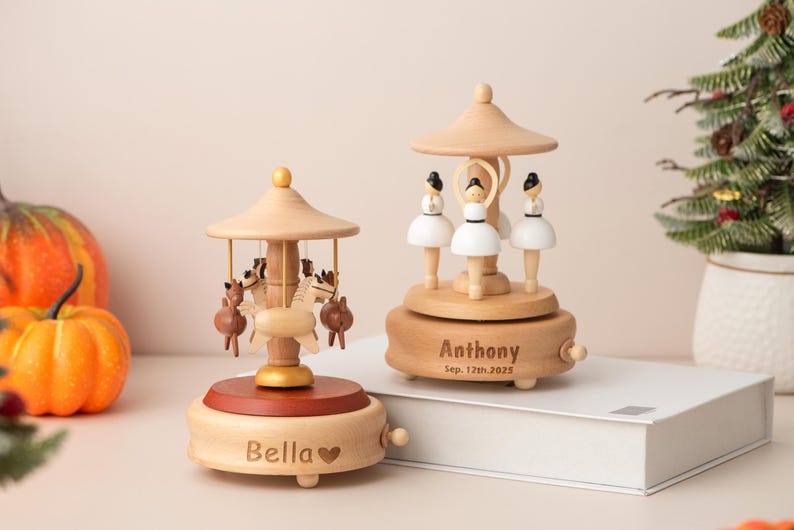 K&ouml;nnte beinhalten: Zwei h&ouml;lzerne Spieluhren mit Karussell-Designs. Eine zeigt Pferde, die andere Ballerinas. Die Spieluhren sind hellbraun mit einem dunkelbraunen Sockel. Eine ist mit "Bella" und die andere mit "Anthony Sep. 12th, 2025" graviert.