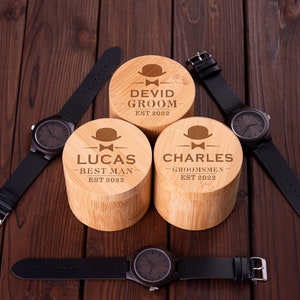 Pu&ograve; includere: Tre scatole di legno con testo inciso e una piccola icona di cappello. Le scatole sono etichettate "David Groom Est 2022", "Lucas Best Man Est 2022" e "Charles Groomsmen Est 2022". Tre cinturini da orologio in pelle nera con quadranti in legno sono posizionati attorno alle scatole.