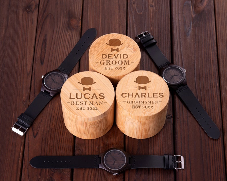 Pu&ograve; includere: Tre scatole di legno con testo inciso e una piccola icona di cappello. Le scatole sono etichettate "David Groom Est 2022", "Lucas Best Man Est 2022" e "Charles Groomsmen Est 2022". Tre cinturini da orologio in pelle nera con quadranti in legno sono posizionati attorno alle scatole.