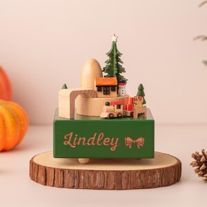 K&ouml;nnte beinhalten: Eine gr&uuml;ne Holzspieluhr mit einer Weihnachtsdorf-Szene, darunter eine Eisenbahn, ein Haus und ein Weihnachtsbaum. Die Box steht auf einem Holzsockel mit dem Namen "Lindley" und einer Schleife. K&uuml;rbisse und Tannenzapfen sind im Hintergrund.