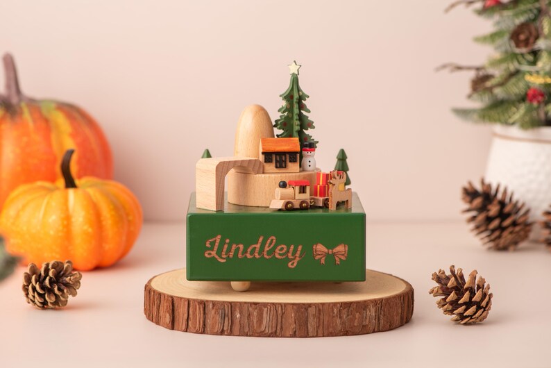 K&ouml;nnte beinhalten: Eine gr&uuml;ne Holzspieluhr mit einer Weihnachtsdorf-Szene, darunter eine Eisenbahn, ein Haus und ein Weihnachtsbaum. Die Box steht auf einem Holzsockel mit dem Namen "Lindley" und einer Schleife. K&uuml;rbisse und Tannenzapfen sind im Hintergrund.