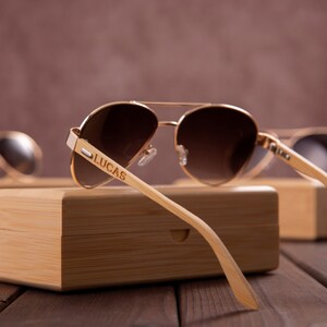 Puede incluir: Un par de gafas de sol aviador doradas con lentes marrones y un marco de madera. Las gafas est&aacute;n apoyadas en una caja de madera con el nombre "LUCAS" grabado en el marco.