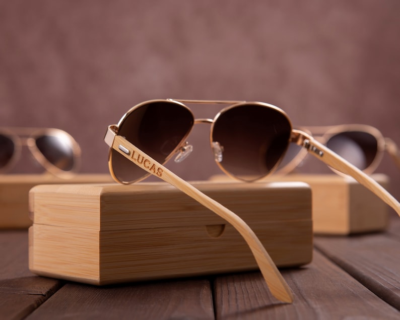 Puede incluir: Un par de gafas de sol aviador doradas con lentes marrones y un marco de madera. Las gafas est&aacute;n apoyadas en una caja de madera con el nombre "LUCAS" grabado en el marco.