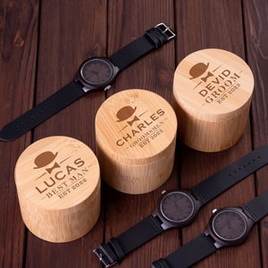 Pu&ograve; includere: Tre scatole di legno con testo inciso e un design a papillon. Le scatole sono etichettate "Lucas Best Man Est 2022", "Charles Groomsmen Est 2022" e "Devid Groom Est 2022".