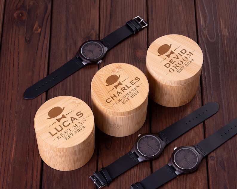 Pu&ograve; includere: Tre scatole di legno con testo inciso e un design a papillon. Le scatole sono etichettate "Lucas Best Man Est 2022", "Charles Groomsmen Est 2022" e "Devid Groom Est 2022".