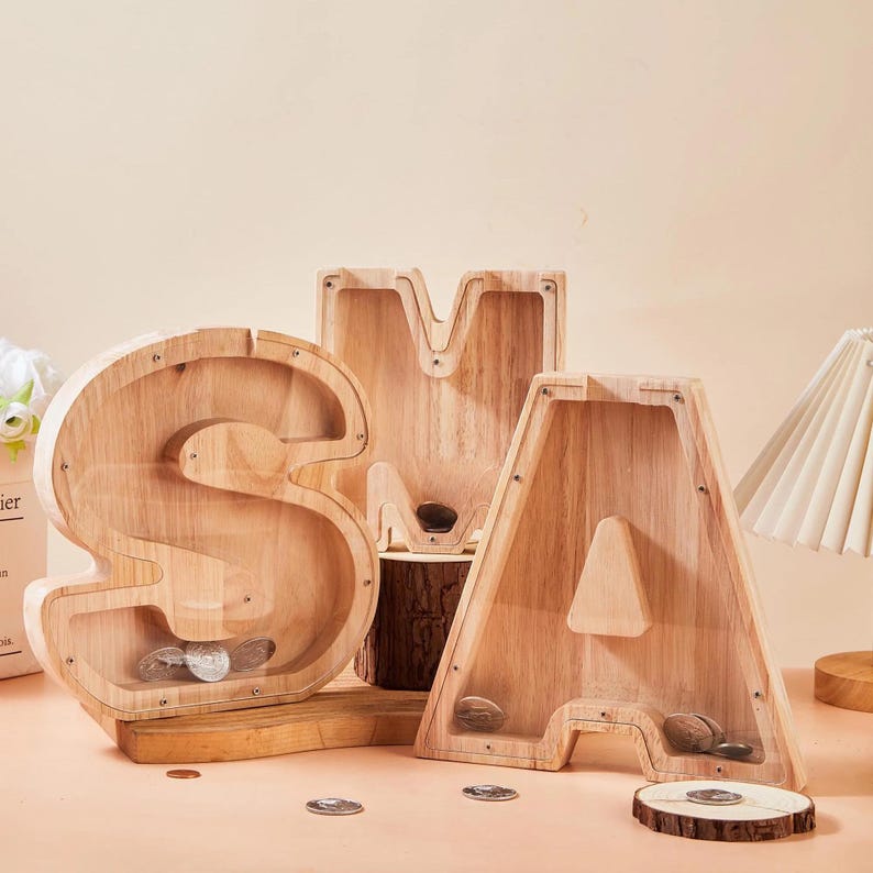 May include: Three wooden letter-shaped money boxes spelling "SAM". The light-coloured wooden banks have clear plastic fronts. The letter "S" is filled with coins. The letter "M" is on a wooden log.