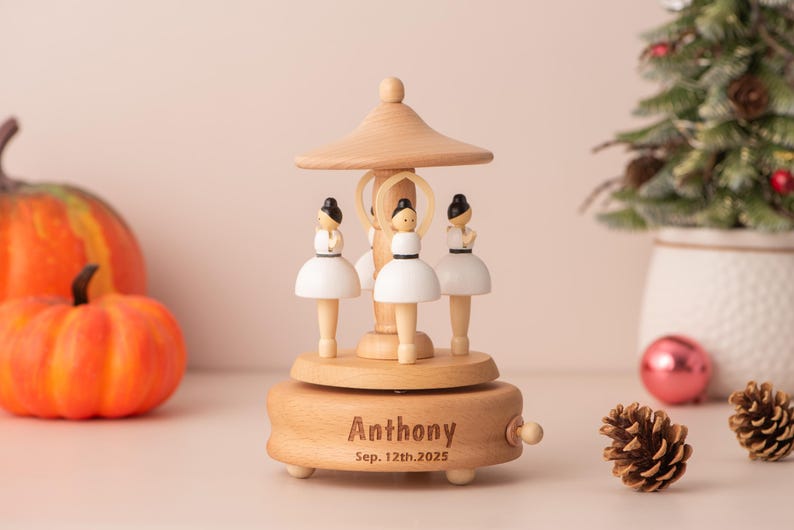 K&ouml;nnte beinhalten: Eine Holzspieluhr mit drei Ballerina-Figuren in wei&szlig;en Tutus und schwarzen Haaren. Auf der Spieluhr sind der Name "Anthony" und das Datum "12. September 2025" eingraviert. Der Hintergrund umfasst K&uuml;rbisse, einen Weihnachtsbaum und Tannenzapfen.