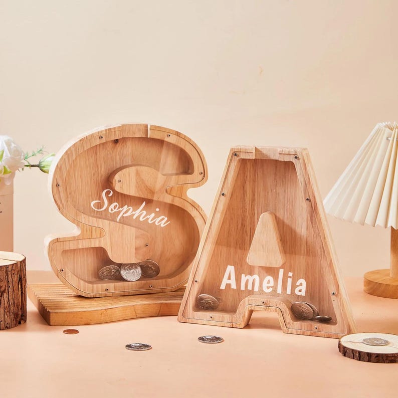 May include: Two wooden letter-shaped money boxes, an "S" and an "A", with clear fronts. The "S" is labelled "Sophia" and the "A" is labelled "Amelia". Both contain coins. The money boxes are on a light-coloured surface.