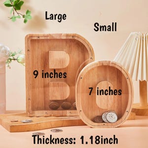 May include: Wooden letter-shaped coin banks, one a "B" and the other a "C". The "B" is 22.86 cm tall, and the "C" is 17.78 cm tall. Both have a thickness of 3 cm. The banks contain coins and are displayed on a wooden surface.