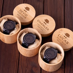 Pu&ograve; includere: Quattro scatole di legno con coperchi incisi, ciascuna contenente un orologio da polso in legno marrone. I coperchi riportano i nomi "Devid Groom Est 2022", "Charles Groomsmen Est 2022", "Lucas Best Man Est 2022", e una quarta scatola con un nome illeggibile.