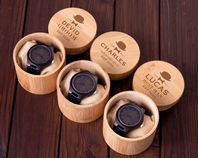 Pu&ograve; includere: Quattro scatole di legno con coperchi incisi, ciascuna contenente un orologio da polso in legno marrone. I coperchi riportano i nomi "Devid Groom Est 2022", "Charles Groomsmen Est 2022", "Lucas Best Man Est 2022", e una quarta scatola con un nome illeggibile.