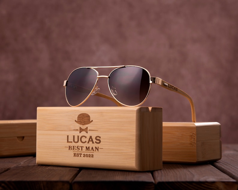 Puede incluir: Un par de gafas de sol aviador marrones con montura dorada y patillas de bamb&uacute;. Las gafas de sol est&aacute;n en una caja de madera con el texto "Lucas Best Man Est 2022" grabado en la parte delantera.