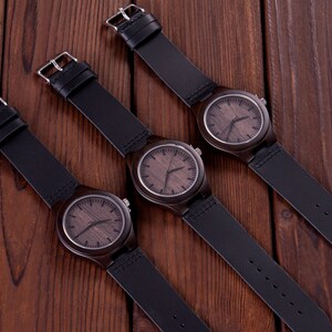 Pu&ograve; includere: Tre orologi da polso in legno con cinturini in pelle nera. Gli orologi hanno quadranti in legno marrone con lancette e indici neri.
