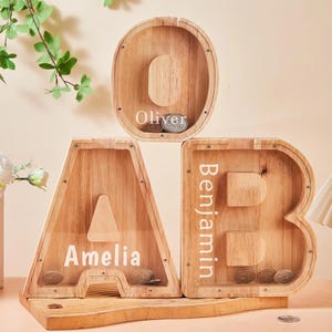 May include: Three wooden piggy banks shaped as the letters A, B, and O. The names "Amelia", "Benjamin", and "Oliver" are printed on the banks. Each bank has a slot for coins. The banks are made of light-coloured wood.