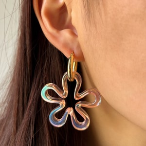 Peut inclure: Une paire de boucles d'oreilles créoles en forme de fleur irisée avec un anneau en métal doré. Les boucles d'oreilles sont en plastique transparent et ont un léger reflet iridescent.