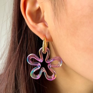 Peut inclure: Une paire de boucles d'oreilles en forme de fleur irisées avec un anneau en or. Les boucles d'oreilles sont en plastique transparent avec un reflet arc-en-ciel.