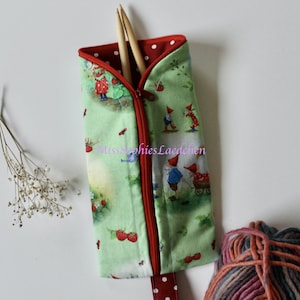 Könnte beinhalten: Eine grüne Reißverschlusstasche mit einem roten, gepunkteten Reißverschluss. Die Tasche ist mit einem Muster aus Gnomen und Erdbeeren verziert. Die Tasche ist offen und zwei hölzerne Stricknadeln befinden sich darin. Der Text "Miss Sophies Laedchen" ist auf die Tasche gedruckt.