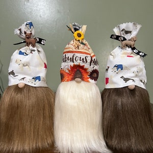 May include: Three gnomes with brown, white, and brown hair. Each gnome is wearing a different fabric hat with a dog design and a small dog figurine on top. The middle gnome's hat has the text "Fabulous Fall" on it.