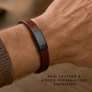 Peut inclure: Un bracelet en cuir marron doté d'un fermoir en métal noir mat. Le texte sur l'image indique : REAL LEATHER & HIDDEN PERSONALIZED ENGRAVING. Le bracelet est porté au poignet, mettant en valeur la texture lisse du cuir et la boucle sombre.