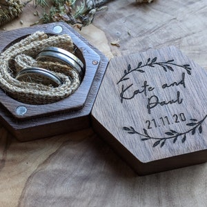May include: A hexagonal wooden ring box with a burlap lining and two silver rings inside. The lid is engraved with the text "Kate and Paul 21.11.20" and a floral design.