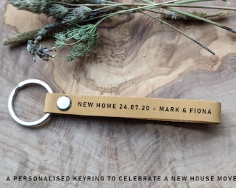 May include: A tan leather keychain with a silver key ring. The keychain has the text "NEW HOME 24.07.20 - MARK & FIONA" printed on it.