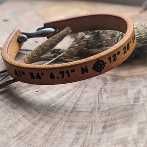 May include: A brown leather bracelet with a compass and coordinates engraved on it. The coordinates are 41° 54' 6.71" N 12° 28' 15"