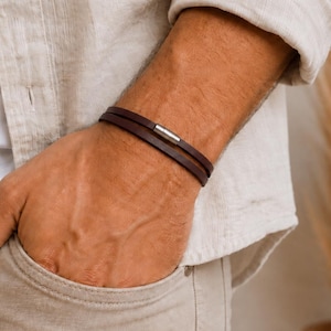 May include: A double-wrap dark brown leather bracelet featuring a slim, rectangular silver-toned metal clasp. The bracelet consists of two thin, smooth leather cords that sit parallel around the wrist, secured by the metallic connector.