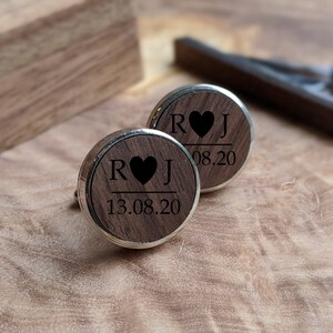 May include: A pair of wooden cufflinks with a silver trim. The cufflinks are engraved with the initials "R" and "J" separated by a heart symbol and the date "13.08.20".